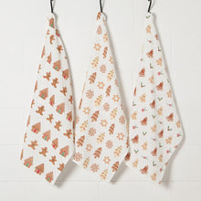Load image into Gallery viewer, Cookie Floursack Towels, three white unbleached cotton dish towels hanging side by side, one printed with brown gingerbread houses and people, another with decorated brown and cream trees, snowflakes, and stars, and a third with brown and cream bells and green and red holly against a white background.