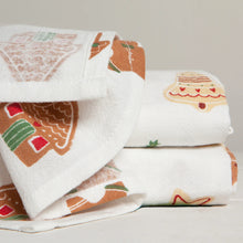 Load image into Gallery viewer, Cookie Floursack Towels, close-up of white unbleached cotton dish towels, with one unfolded towel printed with brown gingerbread houses and people, one folded towel showing a decorated cream bell, and a third folded towel with part of a cream star outlined in red, against a white background. 