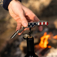 Load image into Gallery viewer, American Flag Double Hinged Bottle Opener, stainless steel double-hinged corkscrew and bottle opener featuring an American flag design, shown opening a wine bottle with a person's hand against a blurred outdoor background with a fire pit.