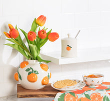 Load image into Gallery viewer, Large Round Vase with Oranges, round white ceramic vase with a glossy finish and raised orange fruit and green leaf accents, displayed with orange tulips on a wooden board beside citrus-themed tableware and food, against a white tiled background.  