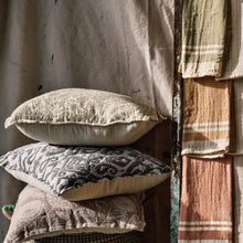 Load image into Gallery viewer, Ikat Jacquard Pillow, lifestyle photo of three stacked pillows with different allover front patterns and natural-colored backs, featuring the gray ikat jacquard design on the centered pillow beside a draped neutral fabric and three hanging striped textiles. 