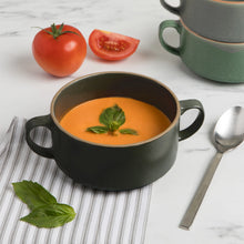 Load image into Gallery viewer, Danica Soup Bowl, Lifestyle scene featuring a matte black stoneware soup bowl with dual side handles filled with tomato soup and garnished with basil leaves, placed next to a metal spoon, fresh basil, and tomatoes, with a stack of matching stoneware bowls in green and gray on a white marble surface. 