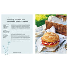 Load image into Gallery viewer, Picnic Cookbook, open cookbook displaying a recipe page beside a photo of a round crusty loaf sandwich filled with sliced red tomatoes, mozzarella, and salami on a white cloth beside a silver knife, a small bowl of green olives, and a brown woven picnic basket holding white plates and glassware, set outdoors on grass, book displayed on white background. 