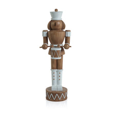 Load image into Gallery viewer, A brown nutcracker figurine holding a drum and drumsticks, featuring a matte finish and white details including a hat, cuffs, boots, and belt. The drum is brown with a white top and bottom and a crisscross design.  The nutcracker stands on a brown base with a white zigzag pattern. Displayed against a white background.