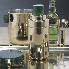 Load image into Gallery viewer, Gold finish metal cocktail shaker with a green resin lid displayed on a reflective tray, with a matching ice bucket with dark green resin handles, a bottle of Chartreuse liqueur, gold finish square cocktail picks, and a stack of dark green round resin coasters in a gold finish holder. Against a dark background.
