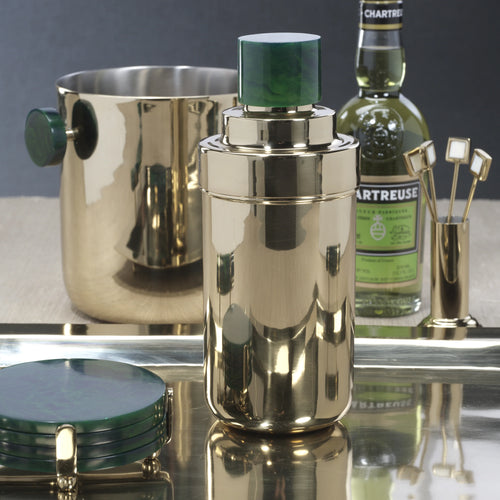 Gold finish metal cocktail shaker with a green resin lid displayed on a reflective tray, with a matching ice bucket with dark green resin handles, a bottle of Chartreuse liqueur, gold finish square cocktail picks, and a stack of dark green round resin coasters in a gold finish holder. Against a dark background.