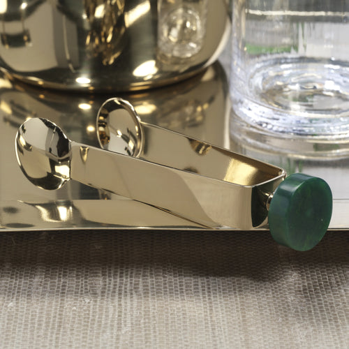 Gold finish metal ice tongs with a round dark green resin accent on the end on a gold finish metal reflective tray. Glass tumbler and partial view of gold finish metal ice bucket also on the tray. On a gray surface.