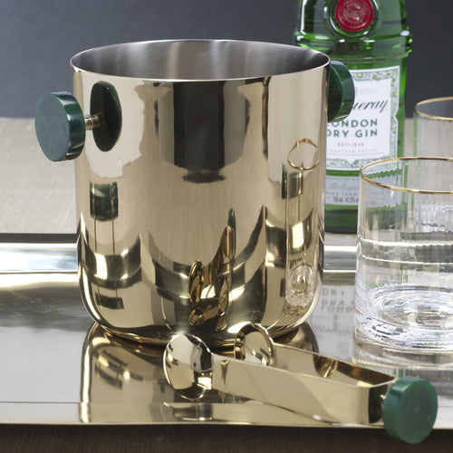 Gold finish ice bucket with round dark green resin handles next to gold finish ice tongs with a round dark green resin knob on the end.  On a reflective metal tray with a glass tumbler with a gold rim. Green bottle of Dry Gin and a glass tumbler with a gold rim behind the ice bucket. Displayed on a white surface and against a dark wall. 