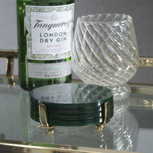 Load image into Gallery viewer, Green glass bottle of Tanqueray London Dry Gin and a clear twisted glass tumbler displayed on a reflective surface with a gold railing. Set of round dark green resin coasters in a gold-tone metal holder with feet in the foreground.