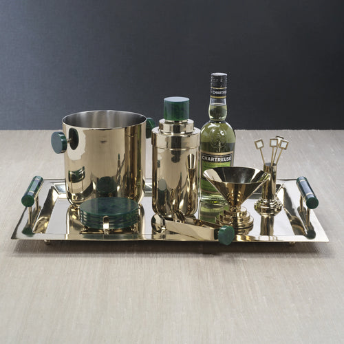 Gold-tone alloy metal bar set on a matching rectangular tray with green resin handles, featuring an ice bucket with green side handles, a cocktail shaker with green lid, a bottle of Chartreuse, a gold funnel-style martini glass, tongs with green handle tips, and gold cocktail picks. All arranged on a light wood surface against a dark background. 