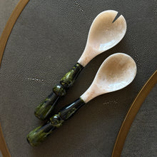 Load image into Gallery viewer, Resin Green Malachite Salad Servers, resin salad server set featuring one serving spoon and one notched serving spoon with marbled ivory heads and glossy dark green handles, lighter green marbling, and sculpted rounded details, displayed on a dark gray textured surface with a gold-tone rim.