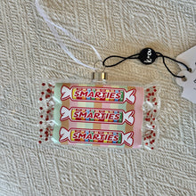 Load image into Gallery viewer, Smarties Candy Ornament, glass  ornament featuring three stacked Smarties candy rolls with transparent wrappers revealing pastel rainbow candies, clear twisted ends accented with red glitter dots, a silver-tone hanging cap, and white ribbon, displayed on cream textured surface.  