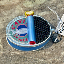 Load image into Gallery viewer, Caviar Ornament, glass ornament shaped like an opened oval tin of caviar with tightly packed rows of raised glossy black caviar pearls, a partially rolled-back silver-blue lid featuring a blue sturgeon illustration and "CAVIAR STURGEON" red text, a gold pull tab, and an iridescent white hanging ribbon, displayed on a gray stone surface. 