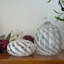 Load image into Gallery viewer, White Swirl Glass Pumpkin, two decorative clear glass pumpkins with white spiral stripes, one short and wide and the other tall, both featuring textured ribbing and clear curved glass stems, displayed on a wooden surface with burgundy and white floral decor and greenery against a white background. 