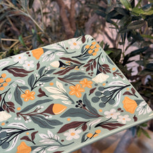 Load image into Gallery viewer, Autumn Acorn Platter, partial view of a rectangle smooth dolomite platter with rounded corners and an acorn, floral, berry, and leafy pattern in shades of orange, brown, white, and gray-green on a pale blue-green background, displayed among greenery. 