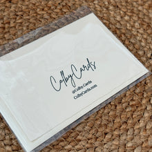 Load image into Gallery viewer, Colby Cards, lifestyle image of the back of a white Lokta paper greeting card in a clear plastic sleeve, featuring the black ColbyCards logo and contact details on a brown woven natural-fiber mat.