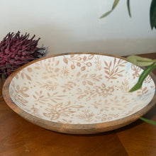 Load image into Gallery viewer, Cream Foliage Shallow Bowl, lifestyle image showing an angled view of a round mango wood bowl with a natural wood rim and cream enamel-coated interior patterned with tan leafy branches, arranged on a wooden table beside dried purple flowers and green foliage.
