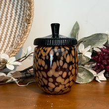 Load image into Gallery viewer, Midnight Pumpkin Glass Acorn Candle, lifestyle image of an acorn-shaped glass candle jar with irregular tan spots on a dark brown background and a glossy black ridged lid with a stem-shaped knob, resting on a wooden surface beside decorative foliage and flowers.
