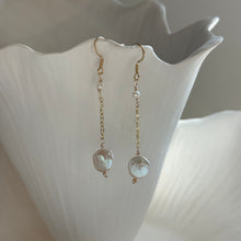 Load image into Gallery viewer, Freshwater Pearl Drop Earrings, angled view of 14k gold-filled earrings with gold-tone curved ear wires, a small white freshwater pearl near the top, and a delicate chain ending in a larger flattened, irregular freshwater pearl, showing the pearls’ lustrous surfaces against a white ridged display.