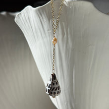 Load image into Gallery viewer, Freshwater Pearl &amp; Spotted Shell Necklace, close-up view of a 14k gold-filled necklace with a main chain forming a V above a small peach-toned freshwater pearl, followed by a short vertical chain drop ending in a white cone shell with dark brown spots, draped over a white ridged display.
