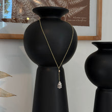 Load image into Gallery viewer, Freshwater Pearl &amp; Spotted Shell Necklace, lifestyle image of a 14k gold-filled necklace with a main chain forming a V above a small freshwater pearl and a short vertical chain drop below the pearl ending in a white cone shell with dark brown spots, displayed around a tall black vase beside a smaller black vase with framed botanical artwork behind them.