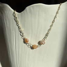 Load image into Gallery viewer, Coral Freshwater Pearl Necklace, close-up view of a 14k gold-filled chain with three rounded, irregular freshwater pearls in peach and pale coral tones, joined by wire-wrapped loops at the center, draped over a white ridged display in direct sunlight.
