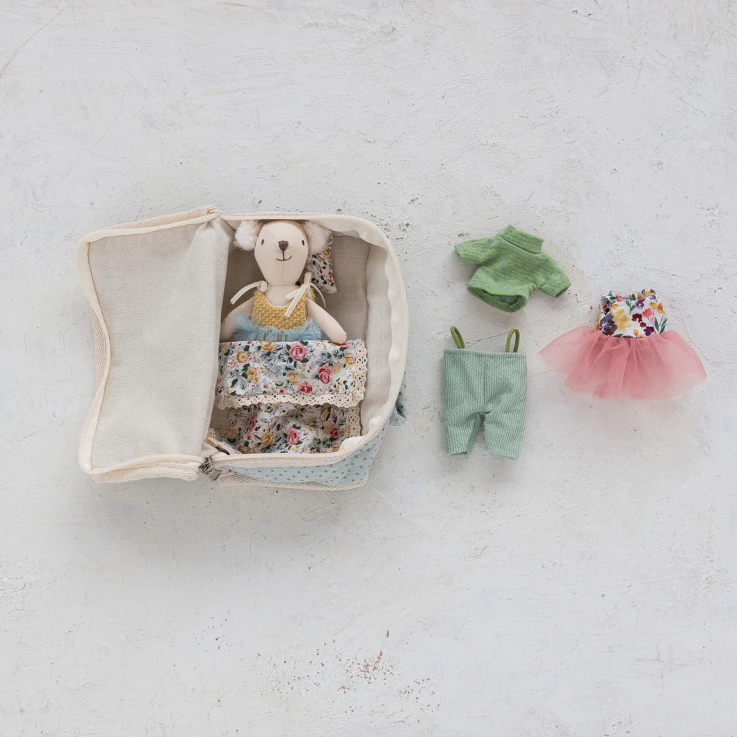 Cream-colored fabric suitcase with floral bedding and pillow, a small plush white bear dressed in a yellow top and blue tulle skirt, shown beside additional outfits including light blue pants, green top, and a floral top with a pink tulle skirt, displayed against a white background.  