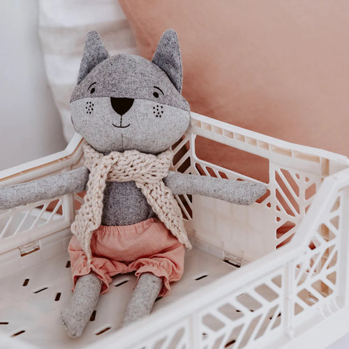 Gray plush raccoon toy wearing blush pink bloomers and a beige Australian merino wool knit scarf, seated inside a cream-colored lattice-style basket against a soft pink background.  