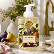Load image into Gallery viewer, Fig &amp; Pear Foaming Soap, lifestyle image of a clear rectangular plastic bottle filled with pale yellow soap and topped with a white foaming pump, featuring an illustrated fig, pear, green leaf, and burgundy ribbon label with a gold-colored seal, displayed beside a bowl of figs and a pear, folded cloths, and a gold-colored faucet.
