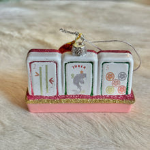 Load image into Gallery viewer, Mahjong Tiles Ornament, lifestyle front view of three white tile shapes on a pink rack with dusty rose pink and gold glitter trim, featuring a two-bamboo design, a fish illustration beneath “JOKER,” and five colored circles, with glitter borders and a silver-colored cap and hanging cord, displayed on a cream fur-textured surface.
