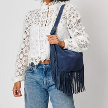 Load image into Gallery viewer, Midnight Reba Hobo, lifestyle image of a model wearing a navy blue suede hobo bag with a curved silhouette, central vertical seam, and long fringe along the sides and bottom, carried on a matching adjustable shoulder strap over a white lace blouse and blue jeans.
