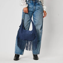 Load image into Gallery viewer, Midnight Reba Hobo, lifestyle image of a model holding a navy blue suede hobo bag by its matching shoulder strap, showing its curved silhouette, central vertical seam, and long fringe along the sides and bottom, styled with a white lace blouse, blue jeans, and black boots, against a white background.
