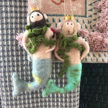 Load image into Gallery viewer, Felt Merman Ornament, two wool felt merman ornaments with gold crowns and green seaweed boas, including one with a dark beard and hair and a pale blue-gray tail accented with yellow and one with a light brown beard and hair and a aqua tail, displayed on patterned textiles. 