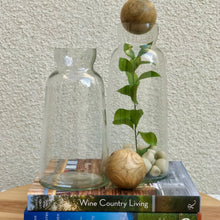 Load image into Gallery viewer, Glass Decanter/ Two Sizes, two clear glass decanters in varying sizes, taller one displayed with a mango wood sphere stopper and filled with decorative stones and green stem, shorter one empty, arranged on stacked books with an additional mango sphere in front, against a textured light-colored wall.
