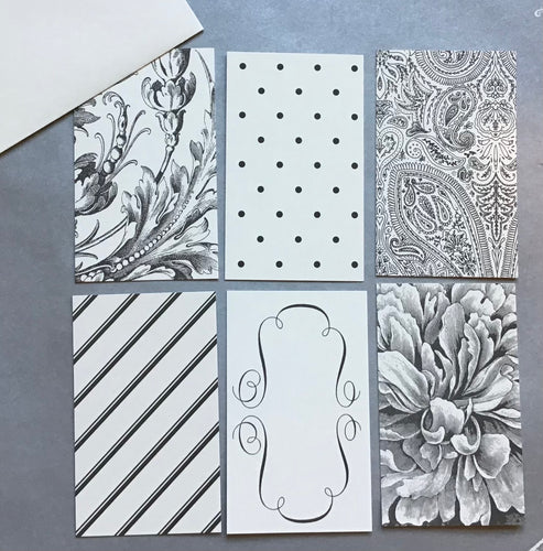 A set of six black and white enclosure cards with different floral and geometric patterns, including a white card with a black floral design, a white card with  black polka dots, a white card with a black paisley design, a white card with black diagonal stripes, a white card with black scrolling curves, and a card with a gray and white flower. A white envelope in the upper left corner on a gray background.