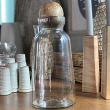 Load image into Gallery viewer, Glass Decanter/ Two Sizes, clear glass decanter with mango wood sphere stopper, displayed on a wood surface alongside white ceramic vessels, woven decor accents, and wood candle holders with gray taper candles. 